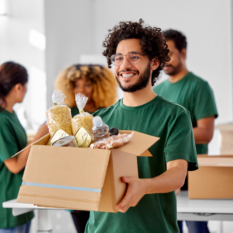 A smiling man in glasses wearing a green -t-shirt offers a box of food with volunteers working in the background.