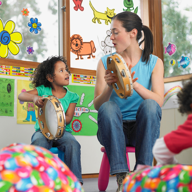 A young girl wearing a green t-shirt and blue jeans and a woman wearing a blue t-shirt and blue jeans sit together in a classroom while playing with tambourines.